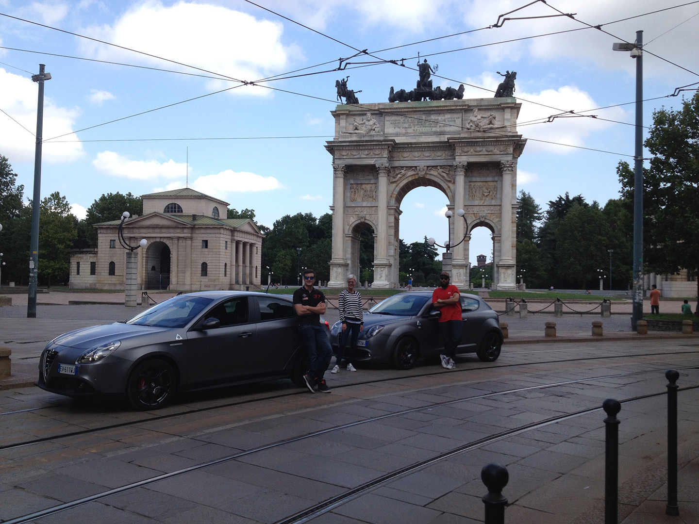 Alfa Romeo Giulietta e MiTo quadrifoglio, grigie, parcheggiate in centro città con a lato i guidatori a braccia conserte che sorridono