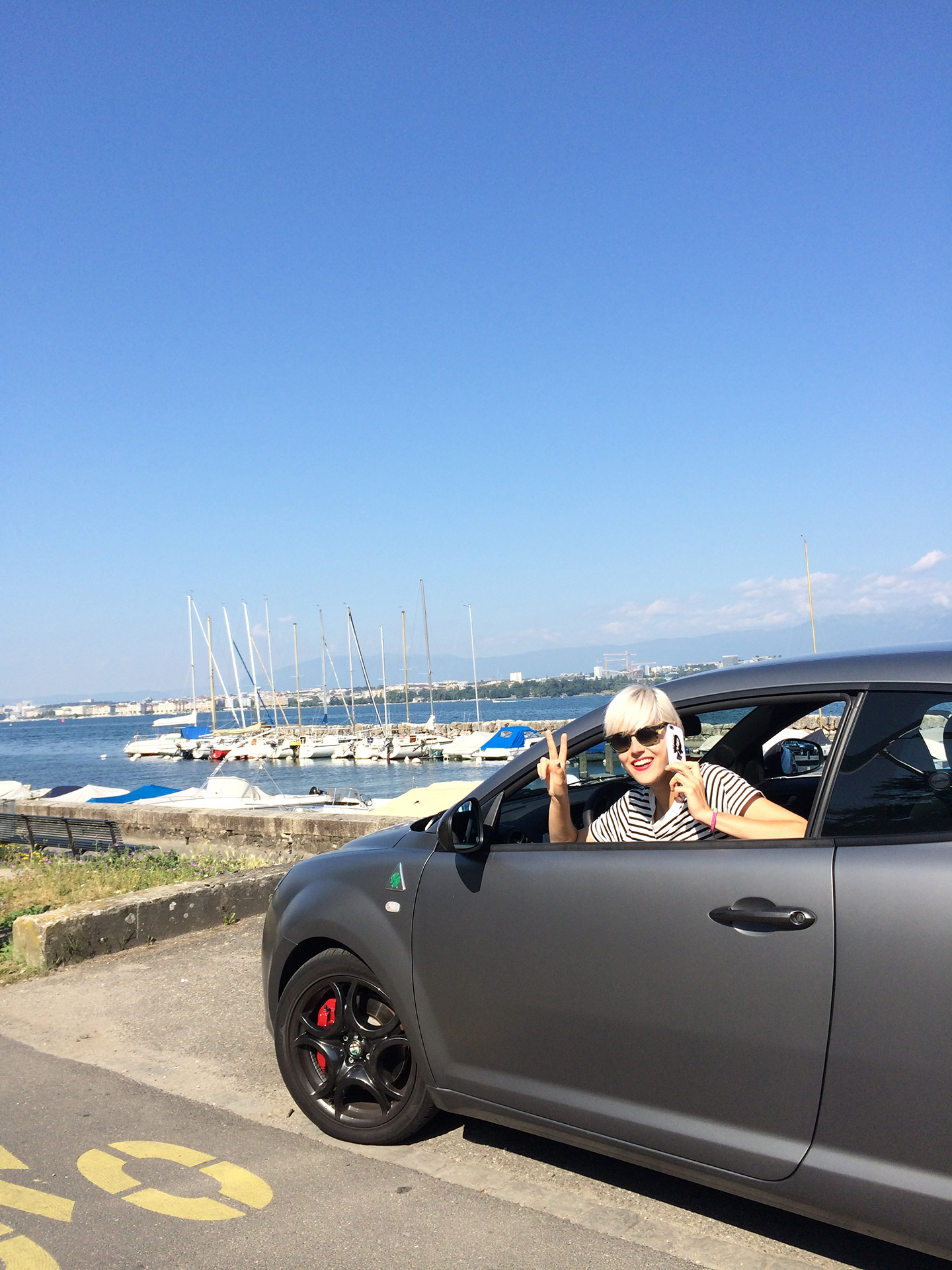 Alfa Romeo MiTo Quadrifoglio grigia con a bordo una ragazza che saluta. Nello sfondo un porto e cielo azzurro