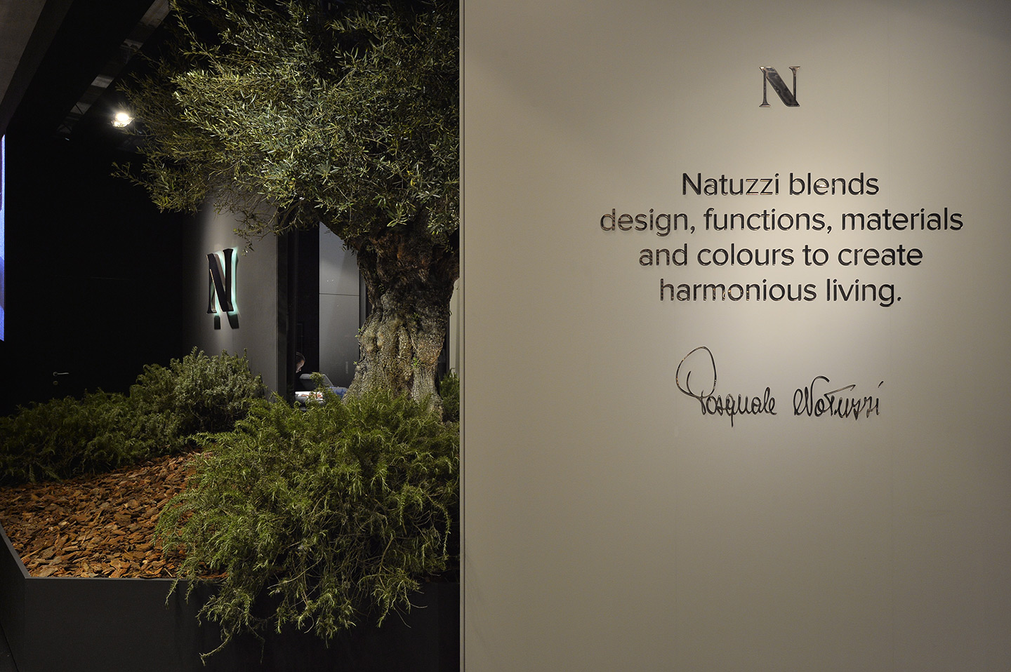 Stand di natuzzi con alberi e cespugli all'ingreso e cartello grigio con scritta: "Natuzzi blends design, functions, materials and colours to create harmonious living." e la firma